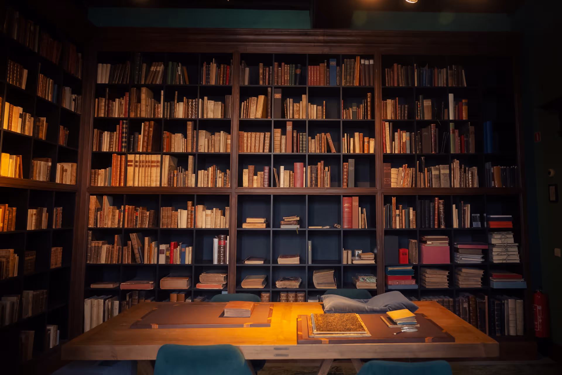 Dimly lit library room with tall bookshelves filled with old books and a wooden table with books and cushions on it.