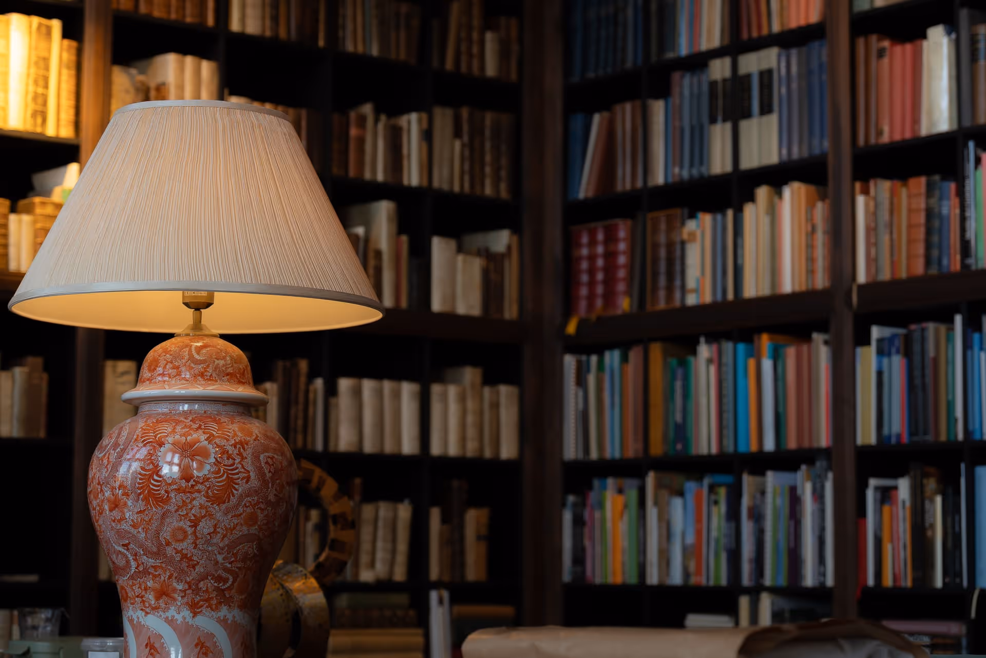 Ornate orange and white ceramic table lamp with pleated cream shade glowing in a cozy room filled with tall bookshelves of various books.