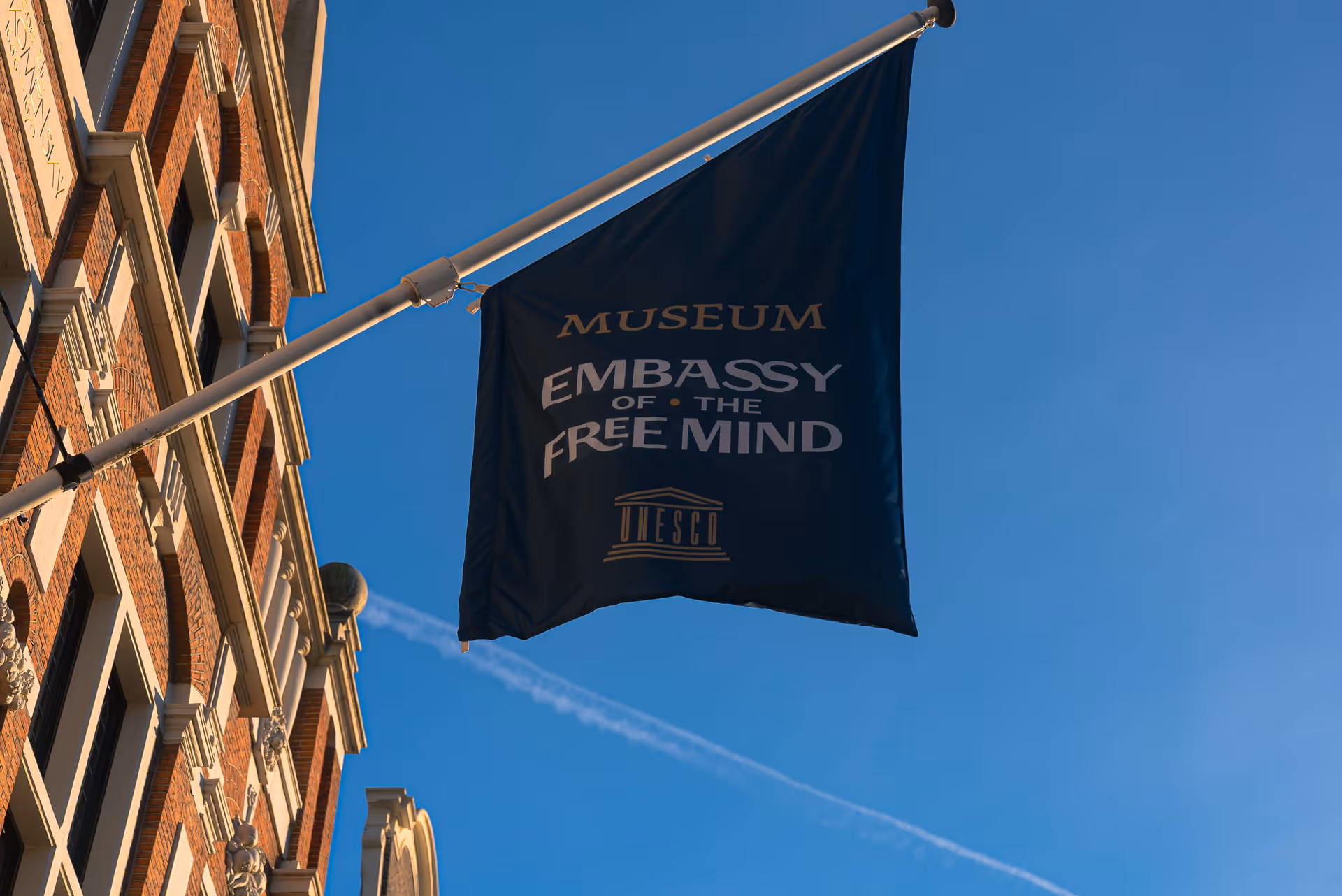 Blue flag with gold and white text reading 'Museum Embassy of the Free Mind UNESCO' hanging from a pole on a building facade against a clear blue sky.