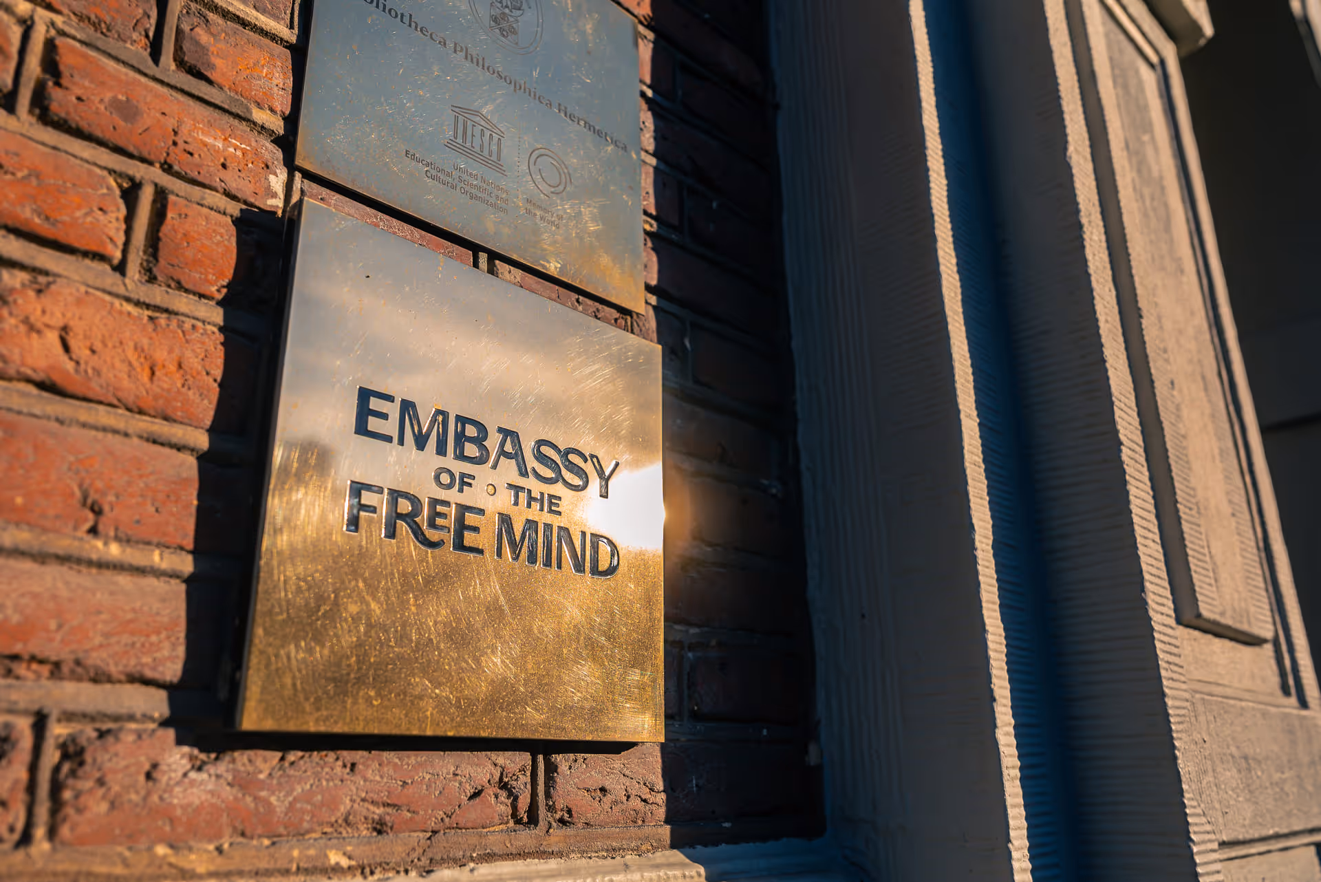 Brass plaque on a brick wall reading 'Embassy of the Free Mind' reflecting sunlight.