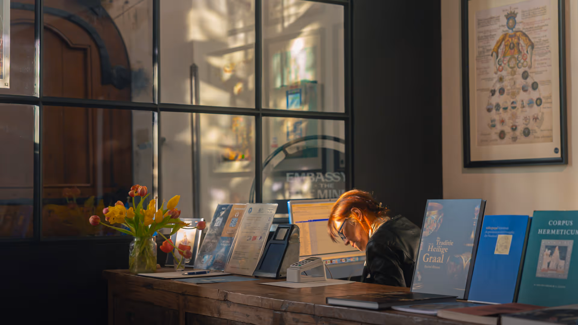 Woman with glasses working at a wooden reception desk with books and a vase of flowers in warm sunlight.