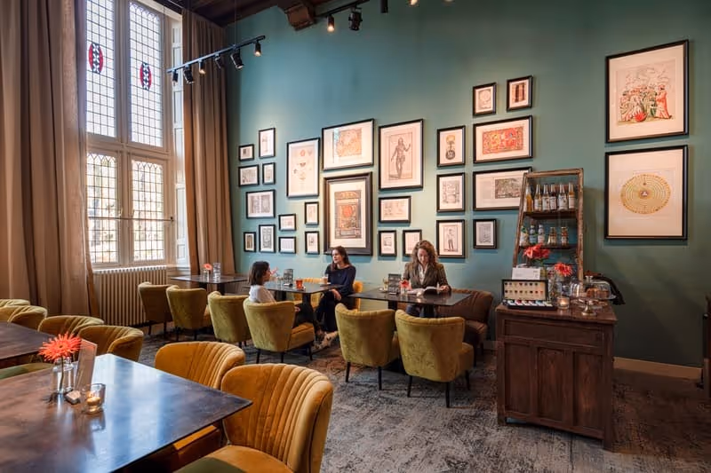 Cafe interior with three people seated at tables against a teal wall adorned with framed vintage artwork and large windows with beige curtains.