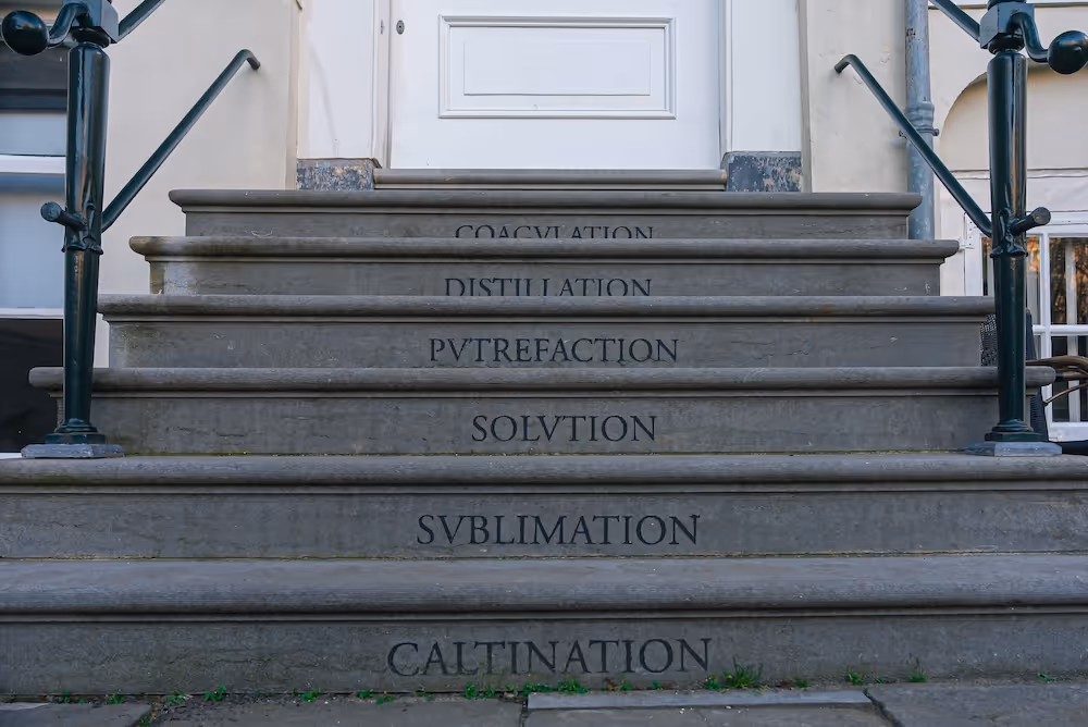 Stone steps with alchemical process words carved on risers leading to a white door.