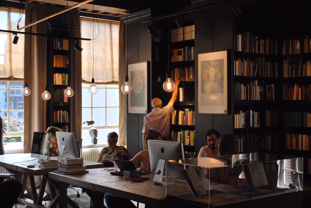 People working and reading in a warmly lit modern library with bookshelves, computers, and hanging bulbs.