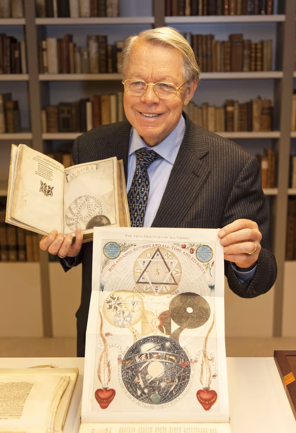 Hij draagt een krijtstreepkostuum en een stropdas met patroon. Achter hem staan planken vol met oude boeken.