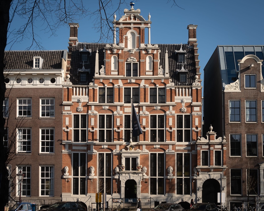 A grand, symmetrical 17th-century canal house with a red brick facade and white stone trim stands under a clear blue sky. Sculpted stone heads adorn the ground floor exterior.