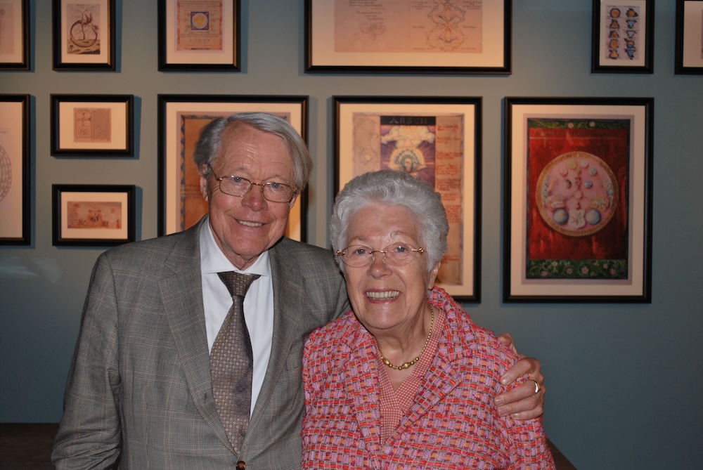 A smiling older man in a grey patterned suit and tie stands next to an older woman with short white hair wearing a pink and white tweed vest. They are posing in front of a wall of framed artwork.