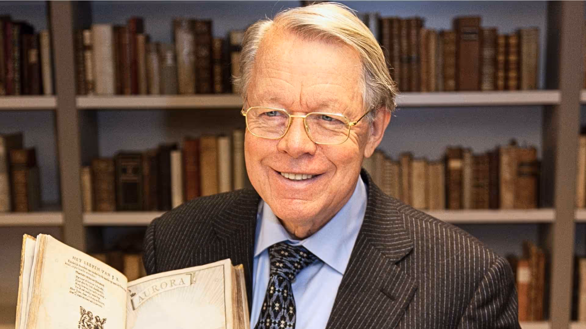 Joost R. Ritman smiles while holding open a rare, historic book. He wears a pinstripe suit in front of a bookshelf.