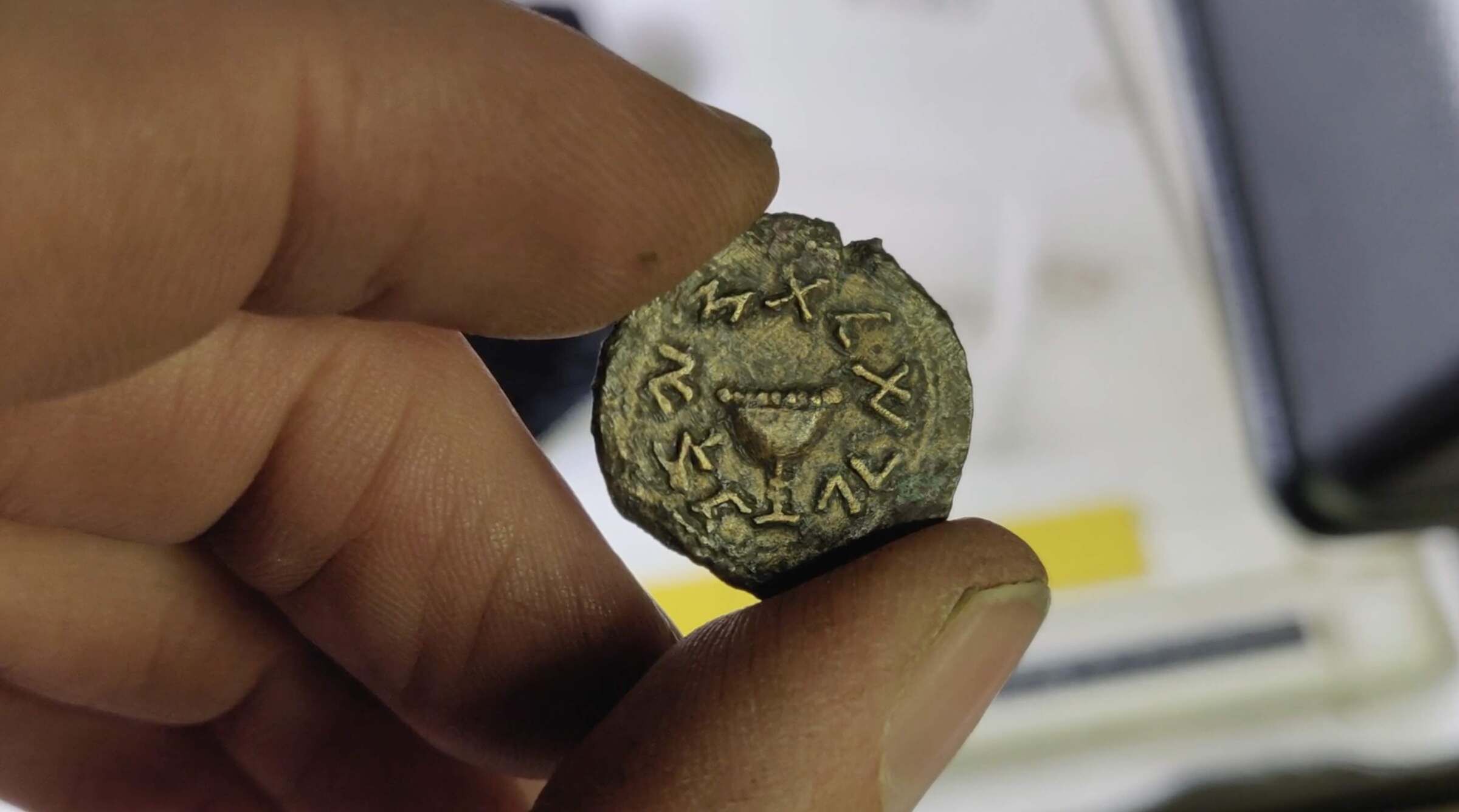A close-up shot of a person's thumb and index finger holding a small, ancient bronze coin.