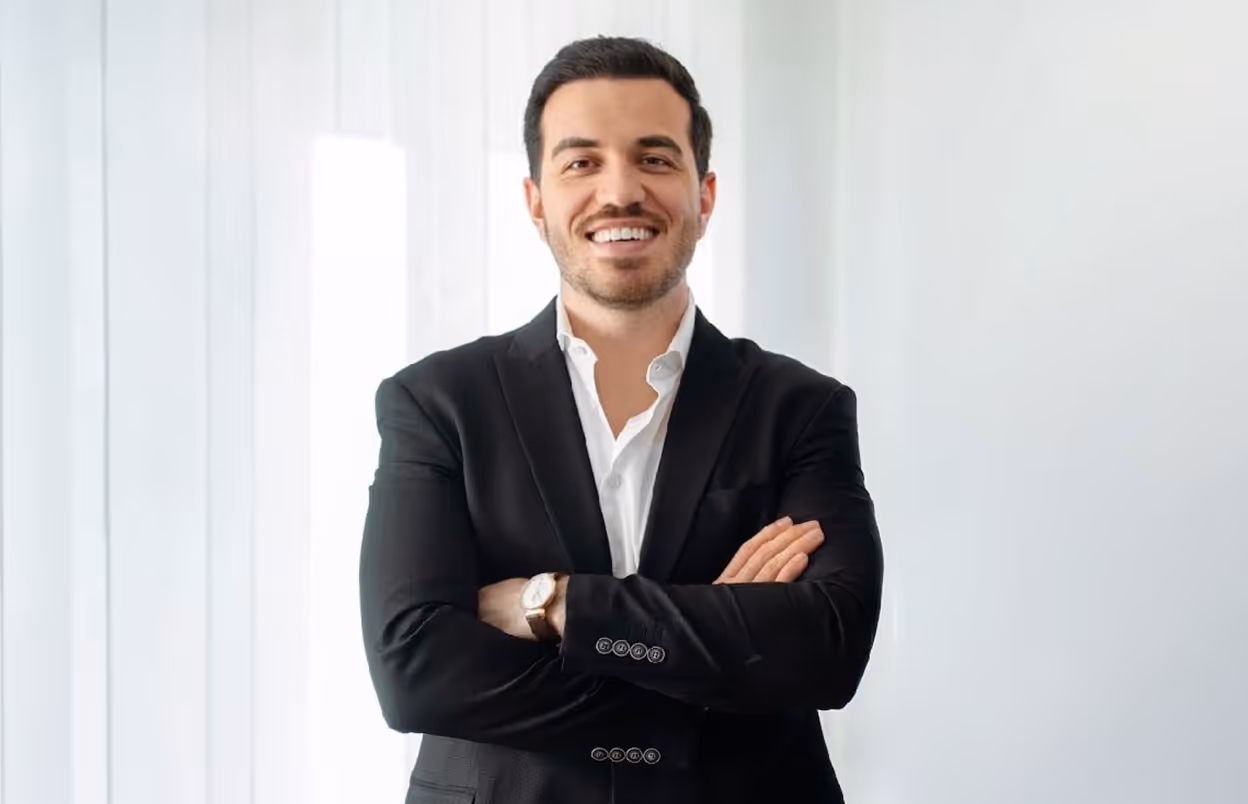 A smiling professional man in a black suit jacket and white shirt, standing with arms crossed against a bright, minimalist background.