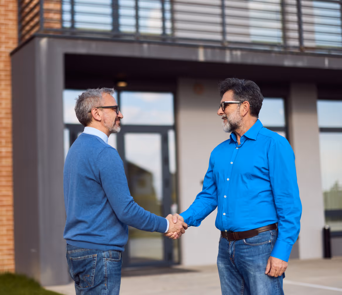 Two middle-aged men with gray hair and beards shaking hands outside a modern building.