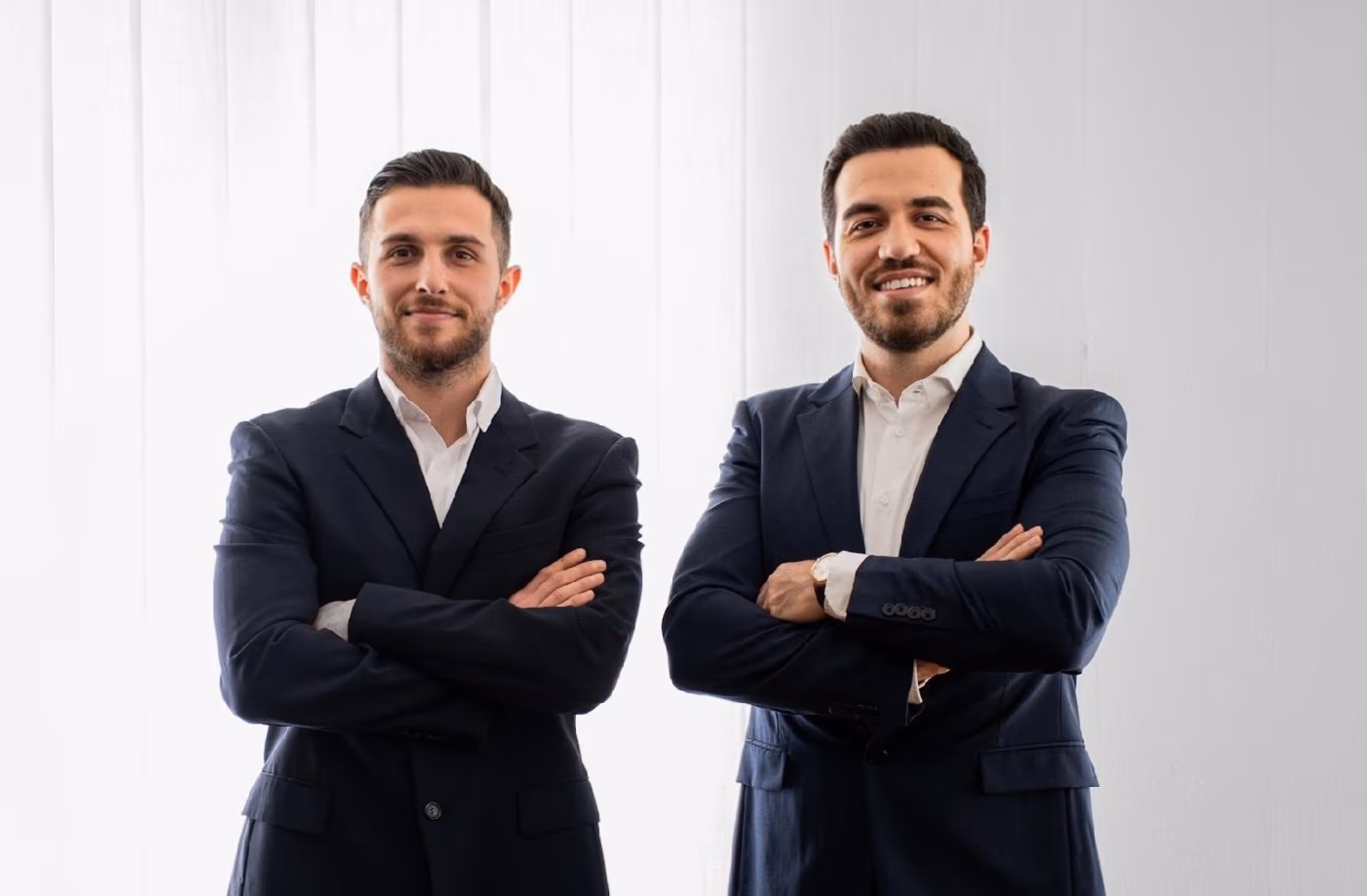 Two smiling men in dark suits with white shirts standing with arms crossed against a bright background.