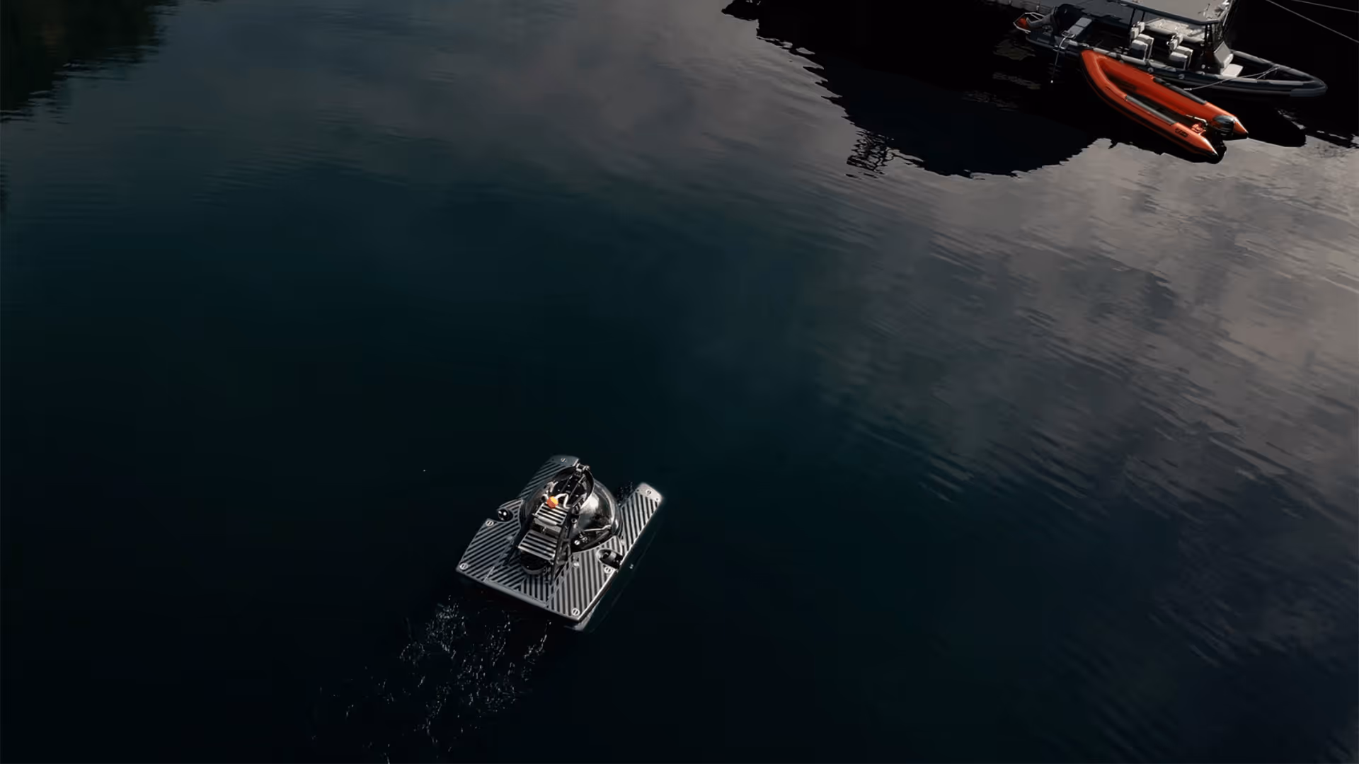 A small, square-shaped subsea remotely operated vehicle (ROV) with a spherical camera or sensor module moves through calm dark water near a dock with two small boats