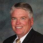 Smiling middle-aged man in a suit and tie with short gray hair against a gray background.