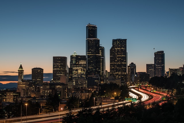 Seattle skyline at dusk with illuminated skyscrapers and light trails from cars on highway.