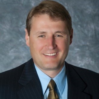 Portrait of a smiling man in a suit, light blue shirt, and gold tie against a gray background.