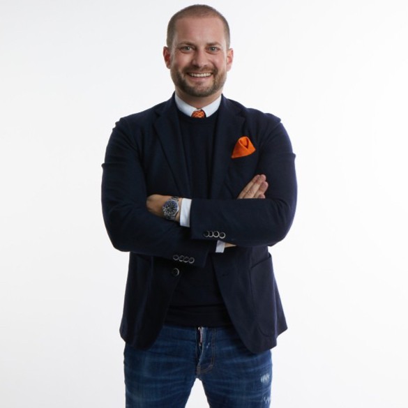 Smiling man with arms crossed wearing a navy blazer, black sweater, jeans, and an orange pocket square.