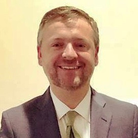 Portrait of a smiling man in a suit, light blue shirt, and gold tie against a gray background.