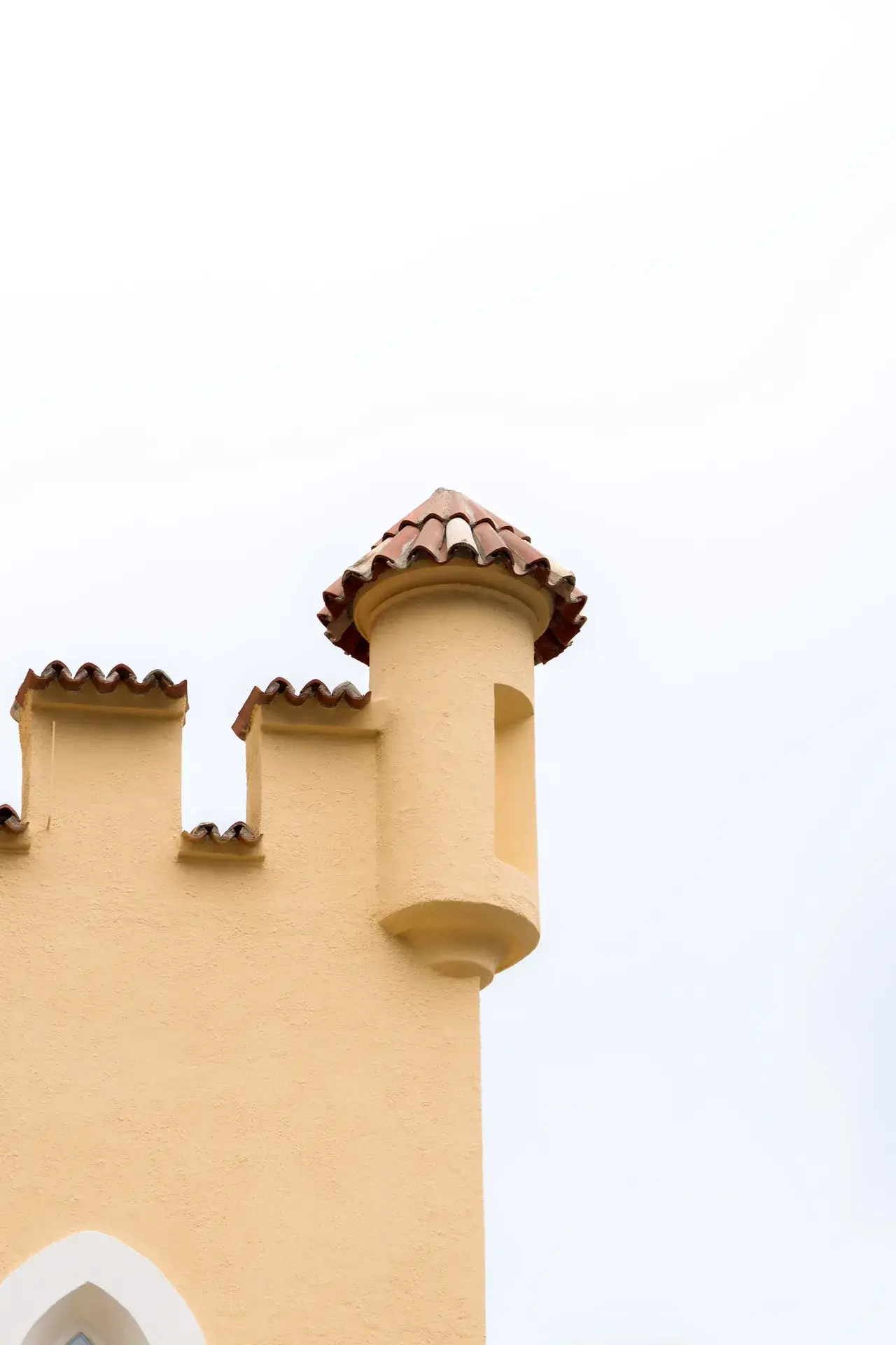 Gelbe Burgmauer mit Zinnen und rundem Türmchen mit rotem Ziegeldach vor weißem Himmel.