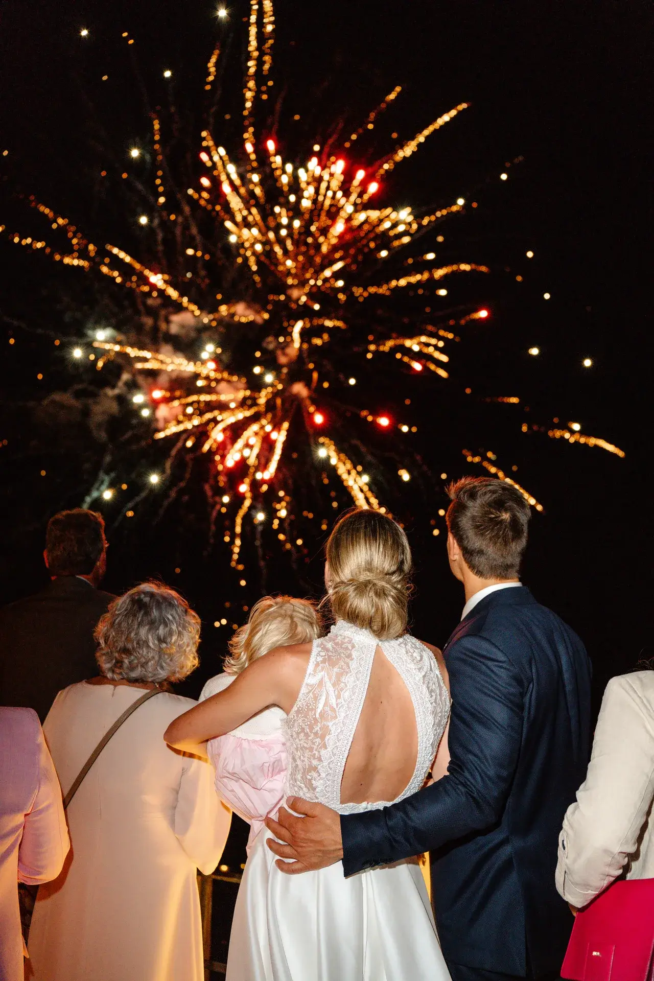 Brautpaar und Gäste schauen bei Nacht ein buntes Feuerwerk an.