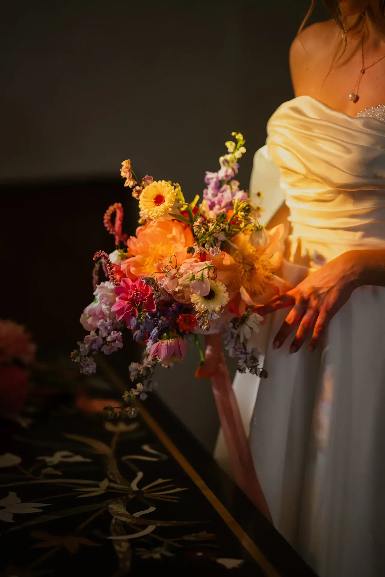 Braut hält einen bunten Blumenstrauß vor dunklem Hintergrund, weiße Kleid und Perlenkette sichtbar.