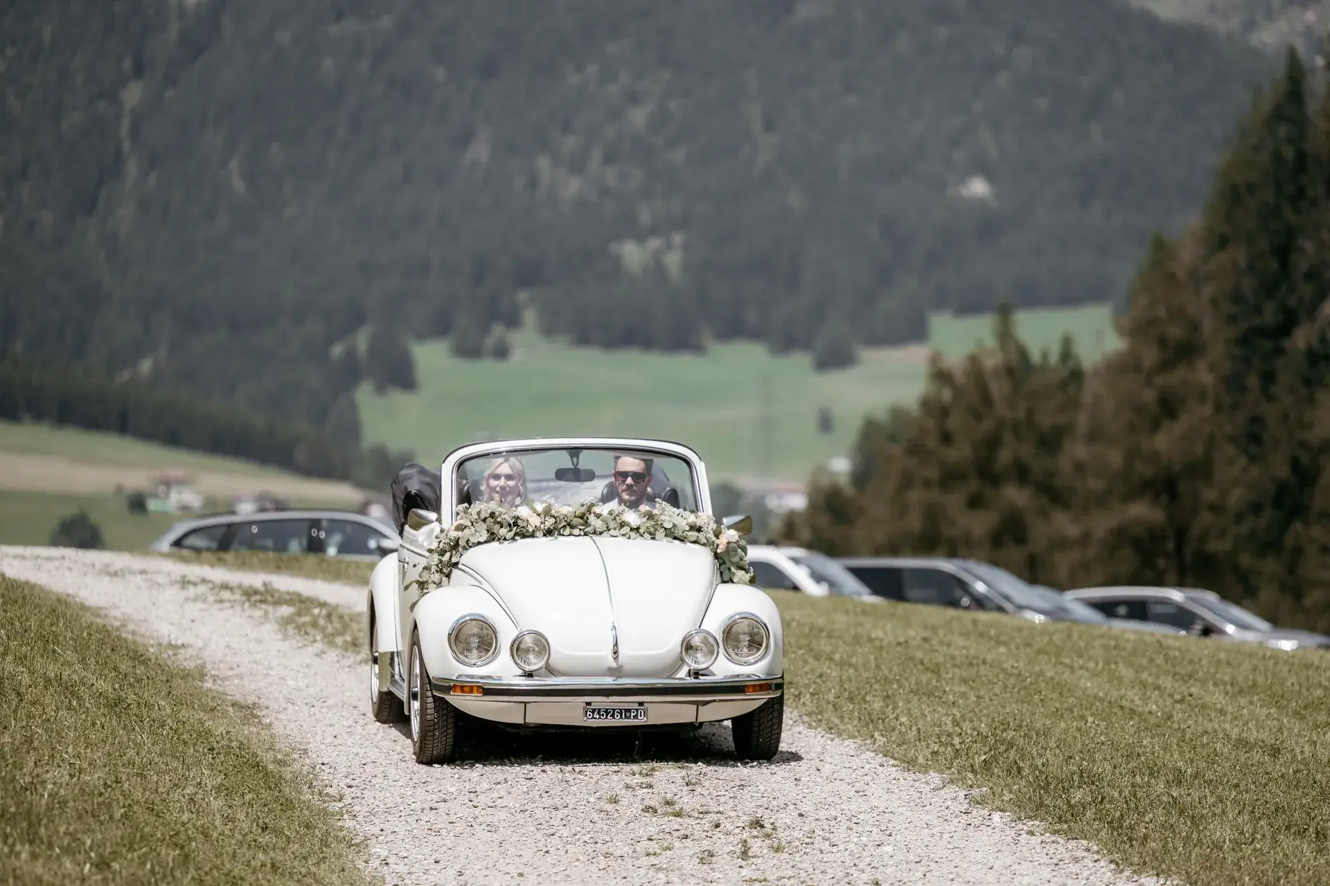 Ein Brautpaar fährt in einem weiß geschmückten Cabrio auf einem Feldweg mit Bergen im Hintergrund.