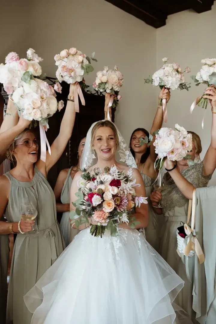 Braut im weißen Kleid mit Brautjungfern, die Rosensträuße hochhalten und lächeln.
