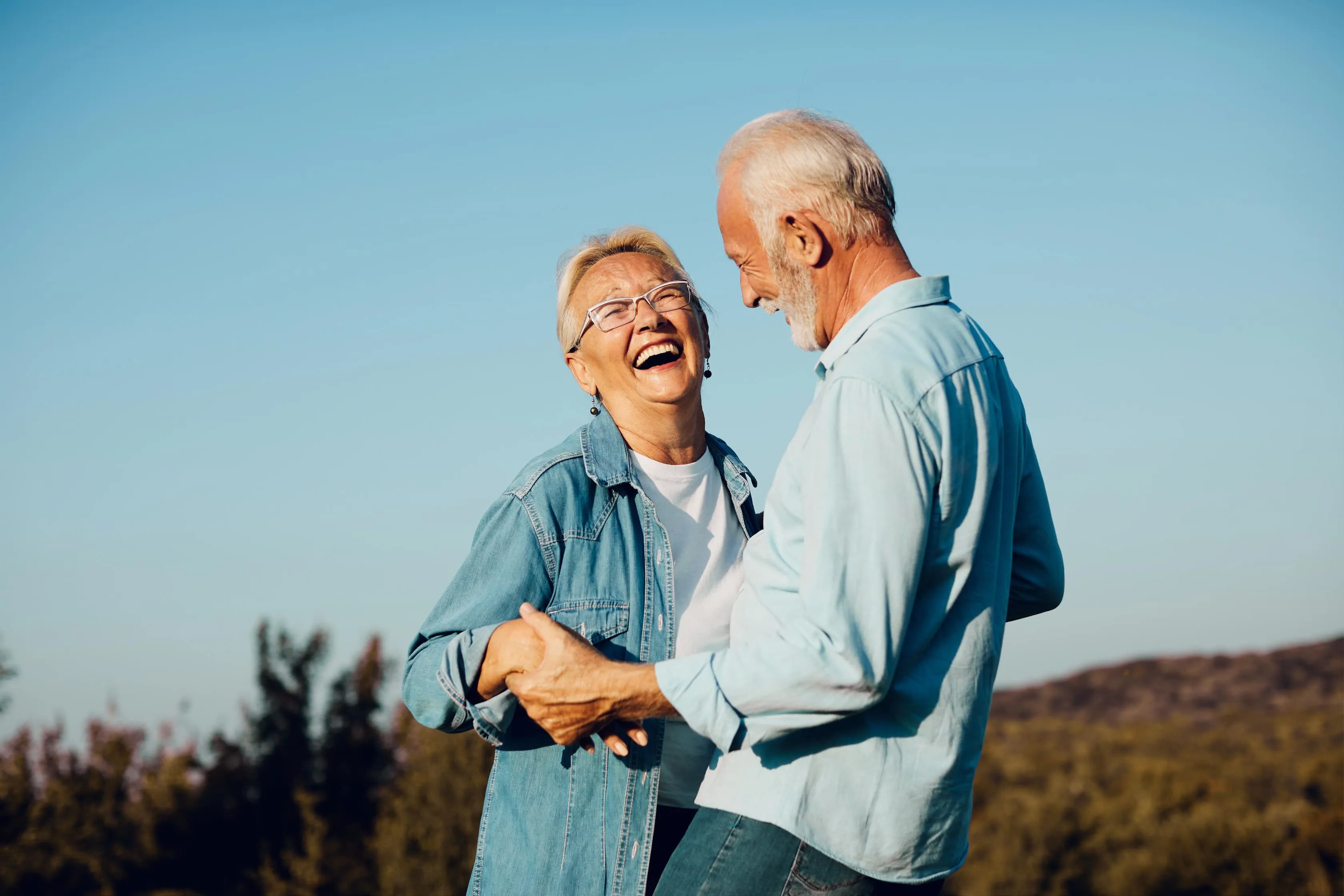 Elderly couple holding hands and laughing outdoors under clear blue sky.