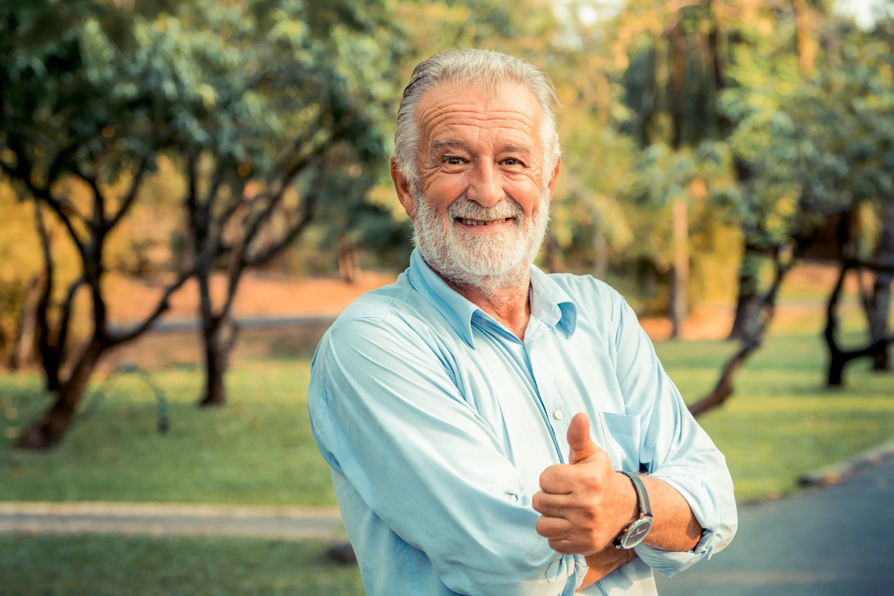 Smiling elderly man with white beard wearing a light blue shirt giving a thumbs up outdoors in a park.