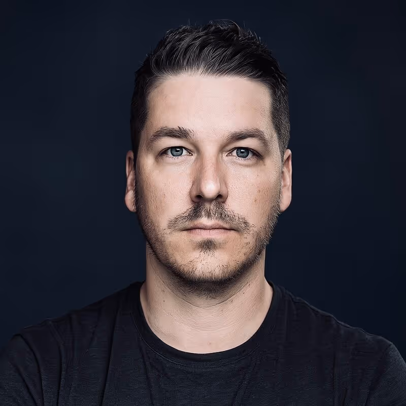 Close-up portrait of a man with short dark hair and light stubble wearing a black shirt against a dark background.