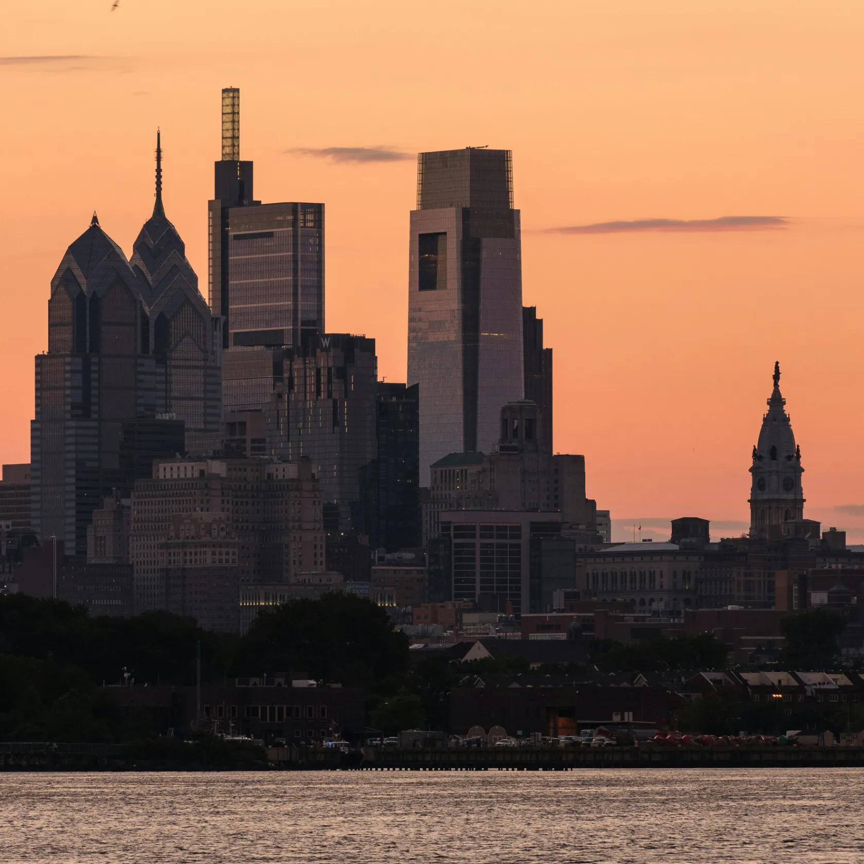 Philadelphia city skyline at sunset with orange sky and Delaware River in the foreground.