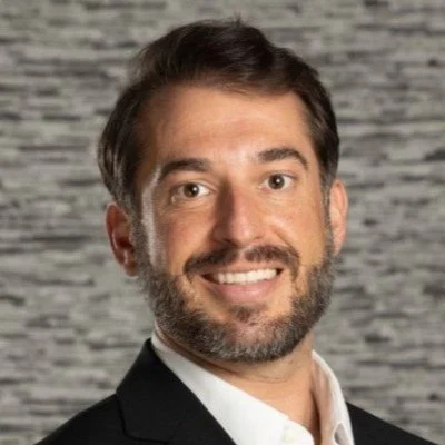 Man with dark hair and beard smiling, wearing a white shirt and black blazer against a blurred gray textured background.