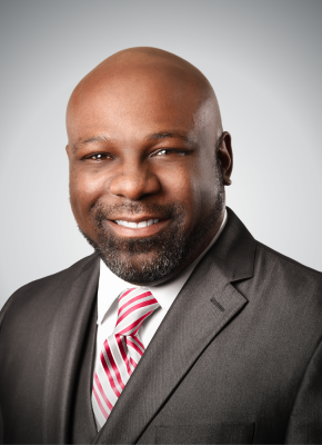 Smiling bald African American man wearing a dark suit and striped red and white tie against a gray background.