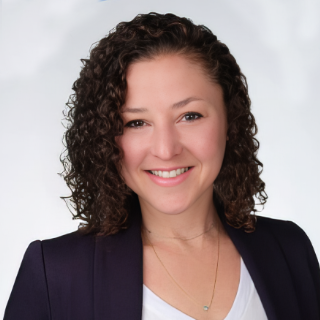 Smiling woman with curly hair wearing a dark blazer and white top on a light background.