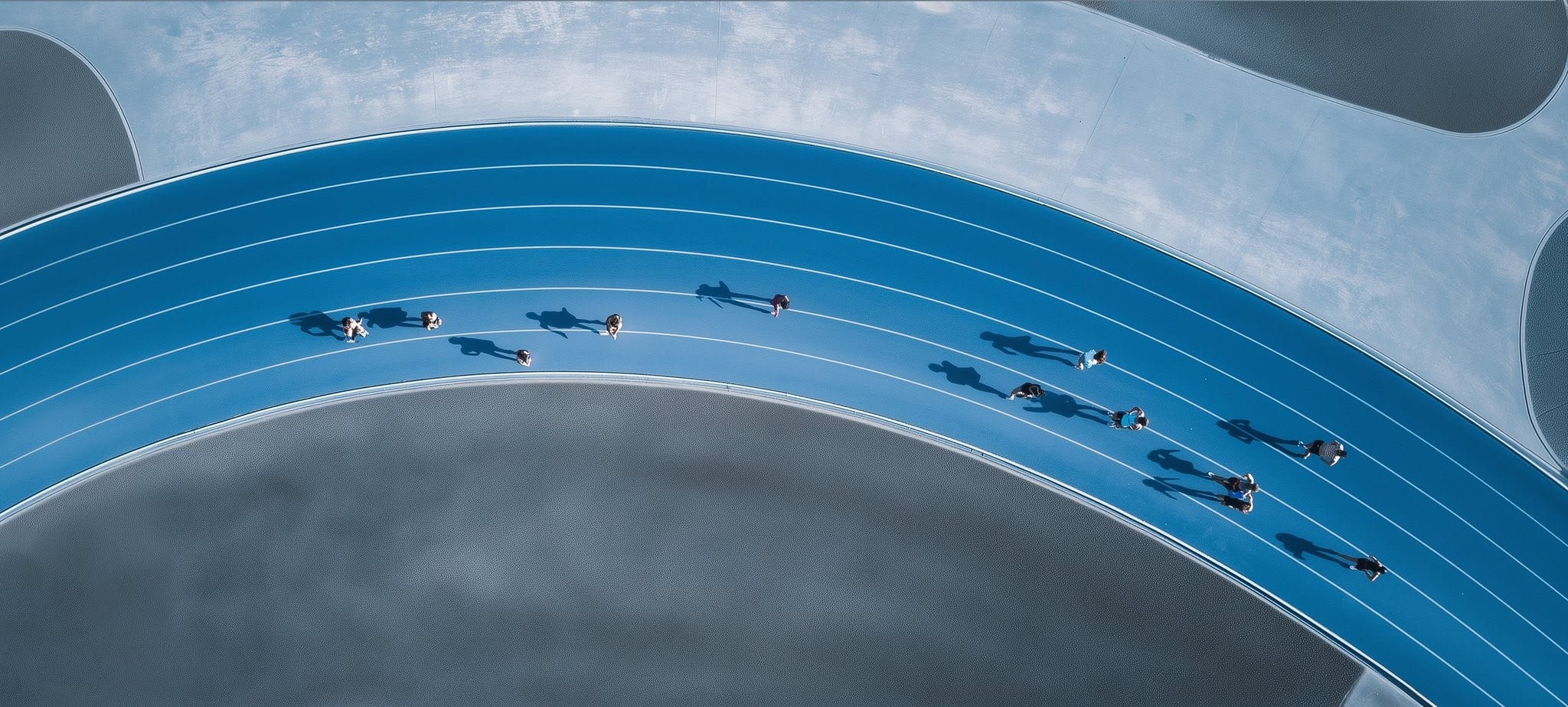Aerial view of people walking and jogging on a curved blue running track casting long shadows.
