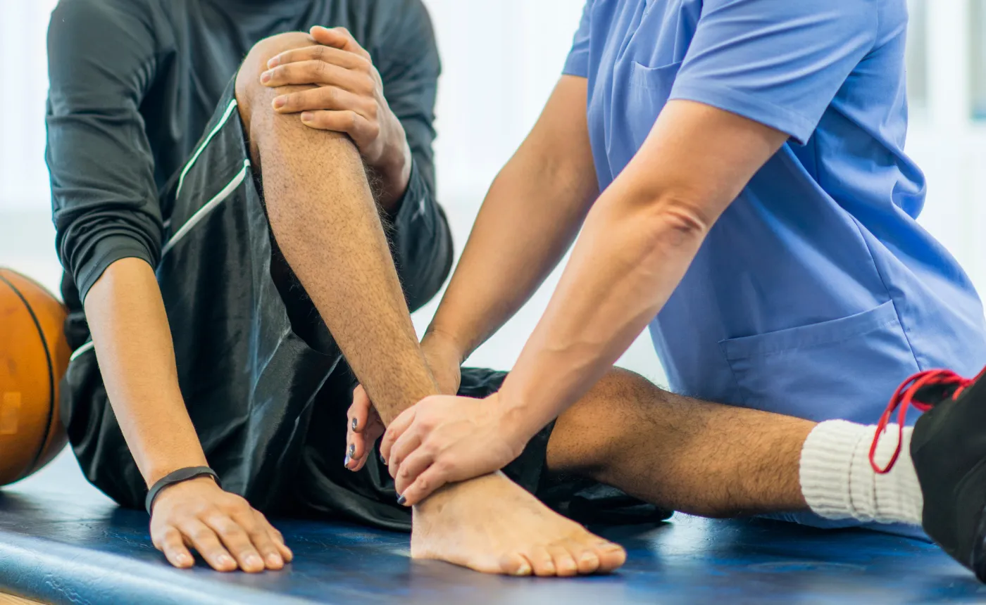Physical therapist in blue scrub examines and supports athlete's ankle during rehabilitation.
