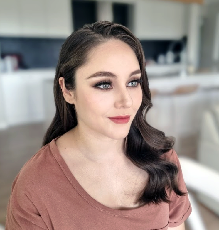 Young woman with light skin, red lipstick, soft brown hair loose with strands framing face, wearing white sheer blouse and gold earrings against plain background.