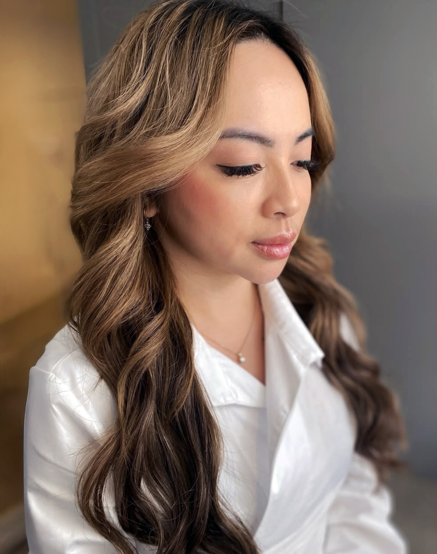 Young woman with light skin, red lipstick, soft brown hair loose with strands framing face, wearing white sheer blouse and gold earrings against plain background.