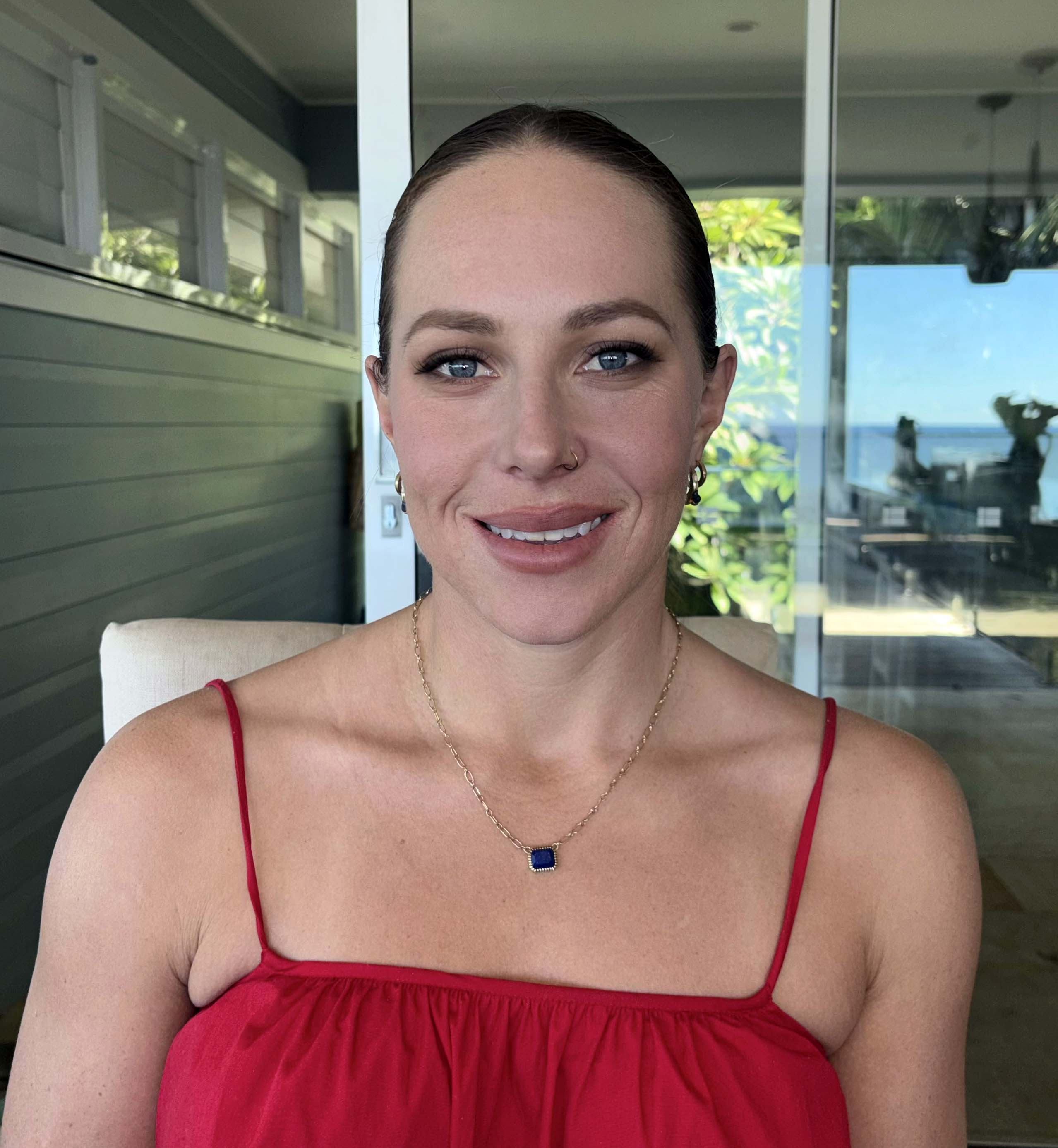 Portrait of a woman with wavy brown hair, bold brown lipstick, and shimmery gold eye makeup wearing an off-shoulder black top.