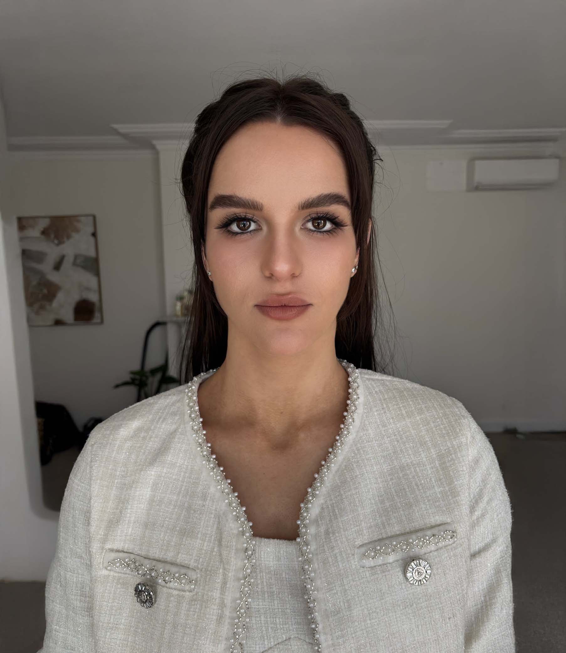 Portrait of a woman with wavy brown hair, bold brown lipstick, and shimmery gold eye makeup wearing an off-shoulder black top.