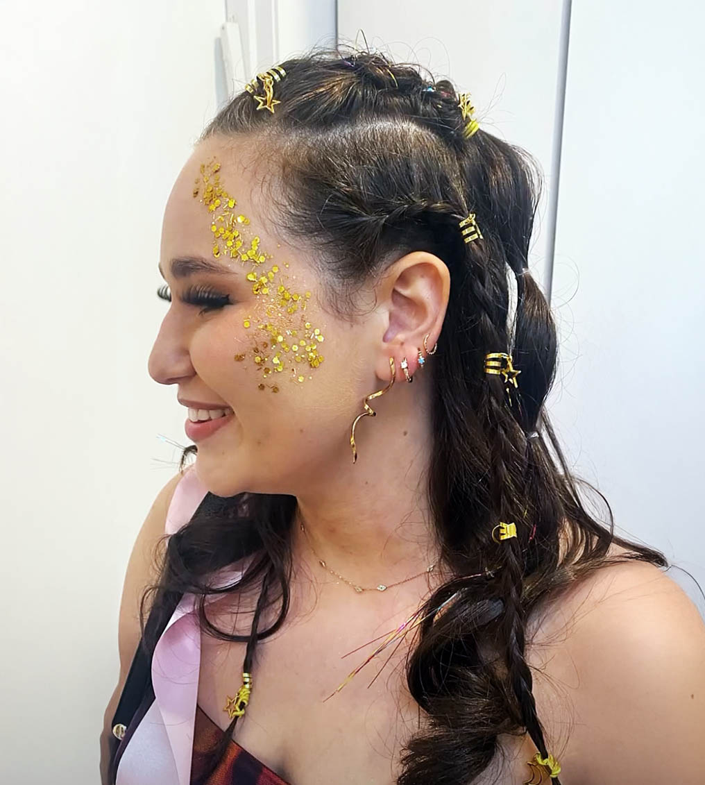 Young woman with vibrant rainbow eye makeup and glossy lips holding a cup filled with paintbrushes, wearing a yellow top.