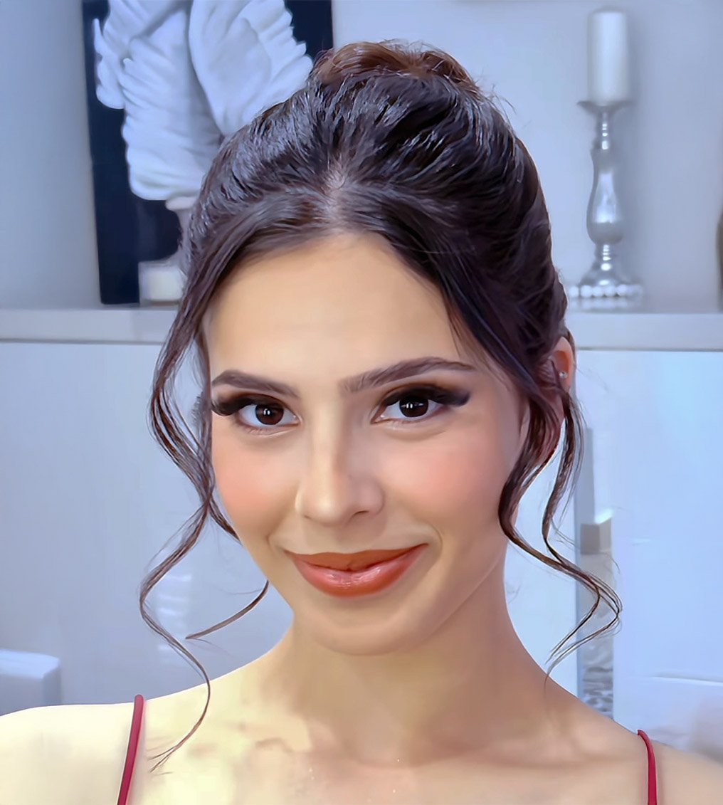 Young woman with vibrant rainbow eye makeup and glossy lips holding a cup filled with paintbrushes, wearing a yellow top.