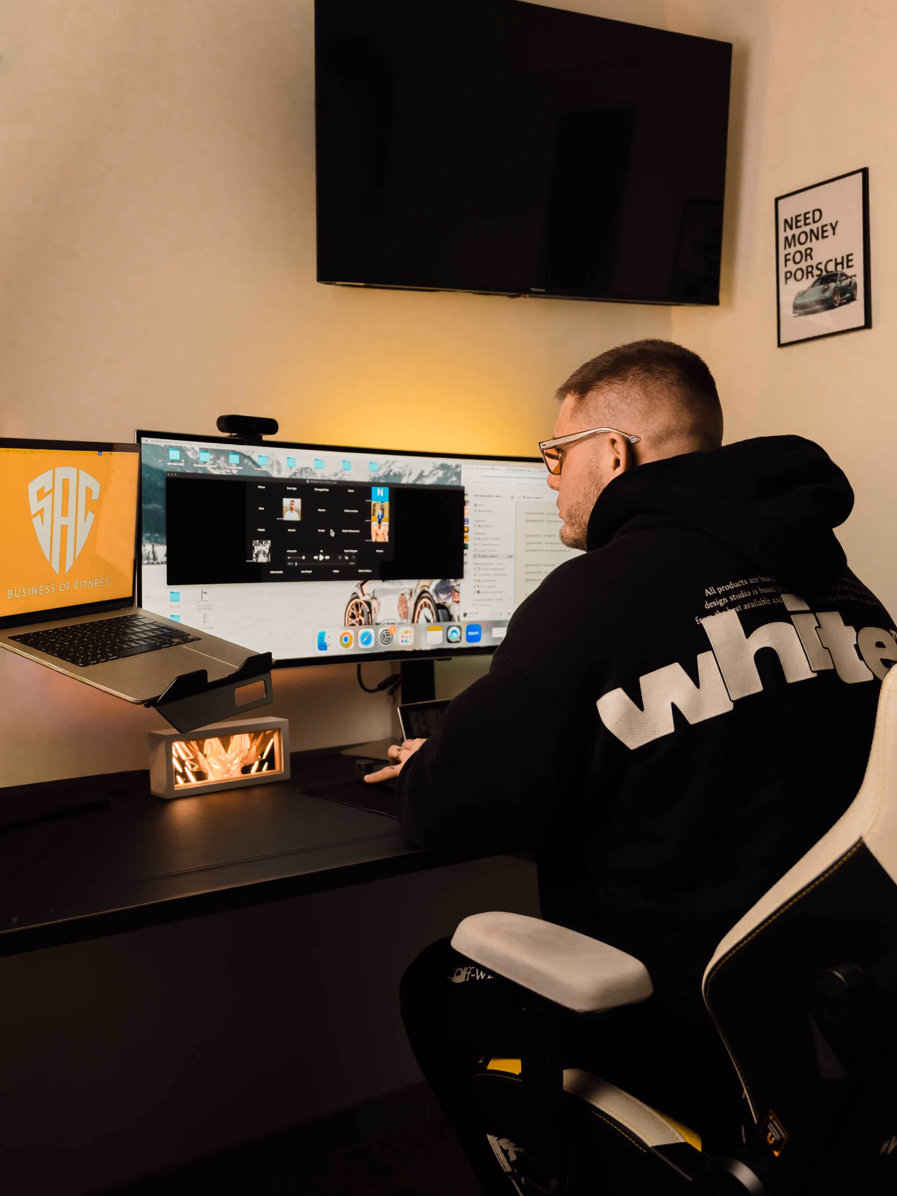 Man wearing glasses and a black hoodie with 'white' text working at a desk with two computer monitors and a laptop.