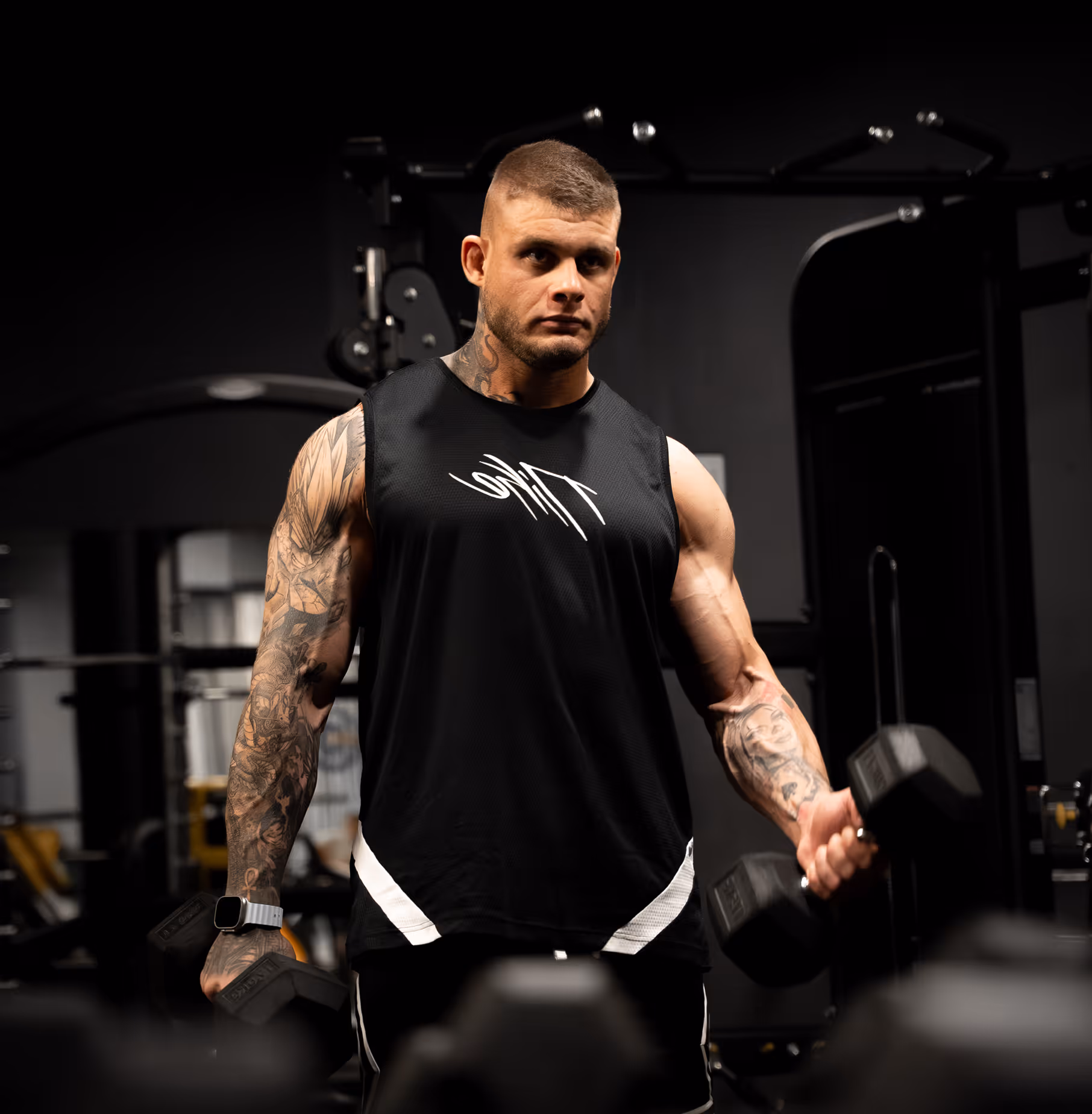 Muscular man with tattoos lifting black dumbbells in a gym.