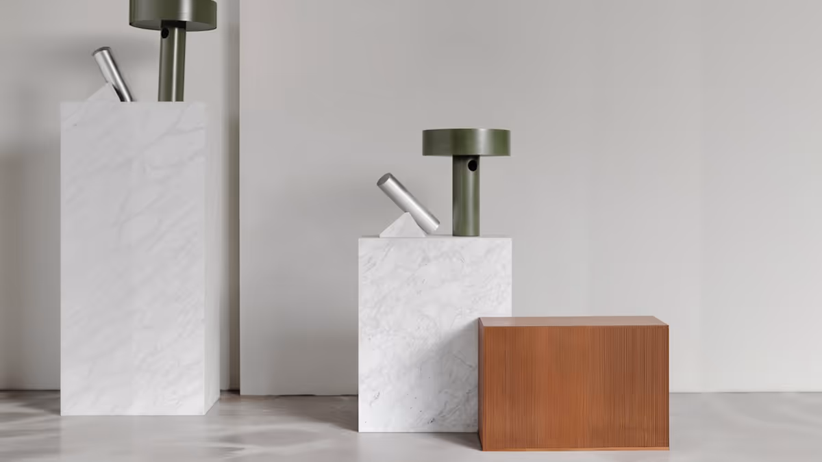 Minimalist still life of two white marble blocks with modern green and silver tubular sculptures, next to a textured brown rectangular block, against a plain light gray wall.