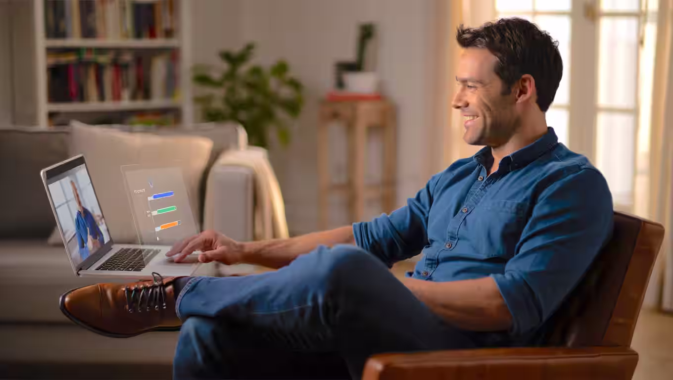 Man in a blue shirt sitting in a chair with legs crossed, smiling and using a laptop to have a call with a custom video course platform developer.