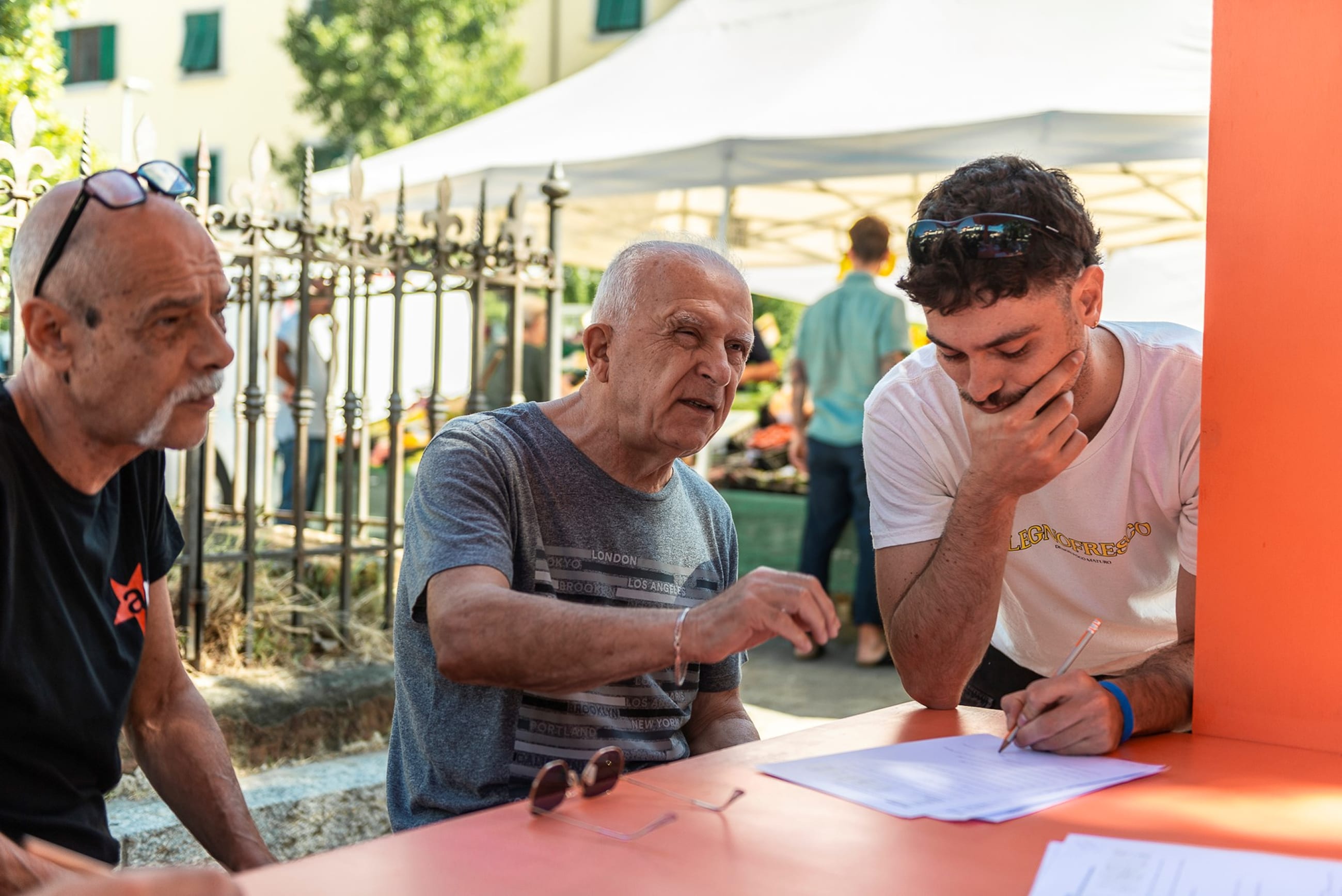 Tre persone compilano dei fogli a un tavolo arancione, con un mercato sullo sfondo