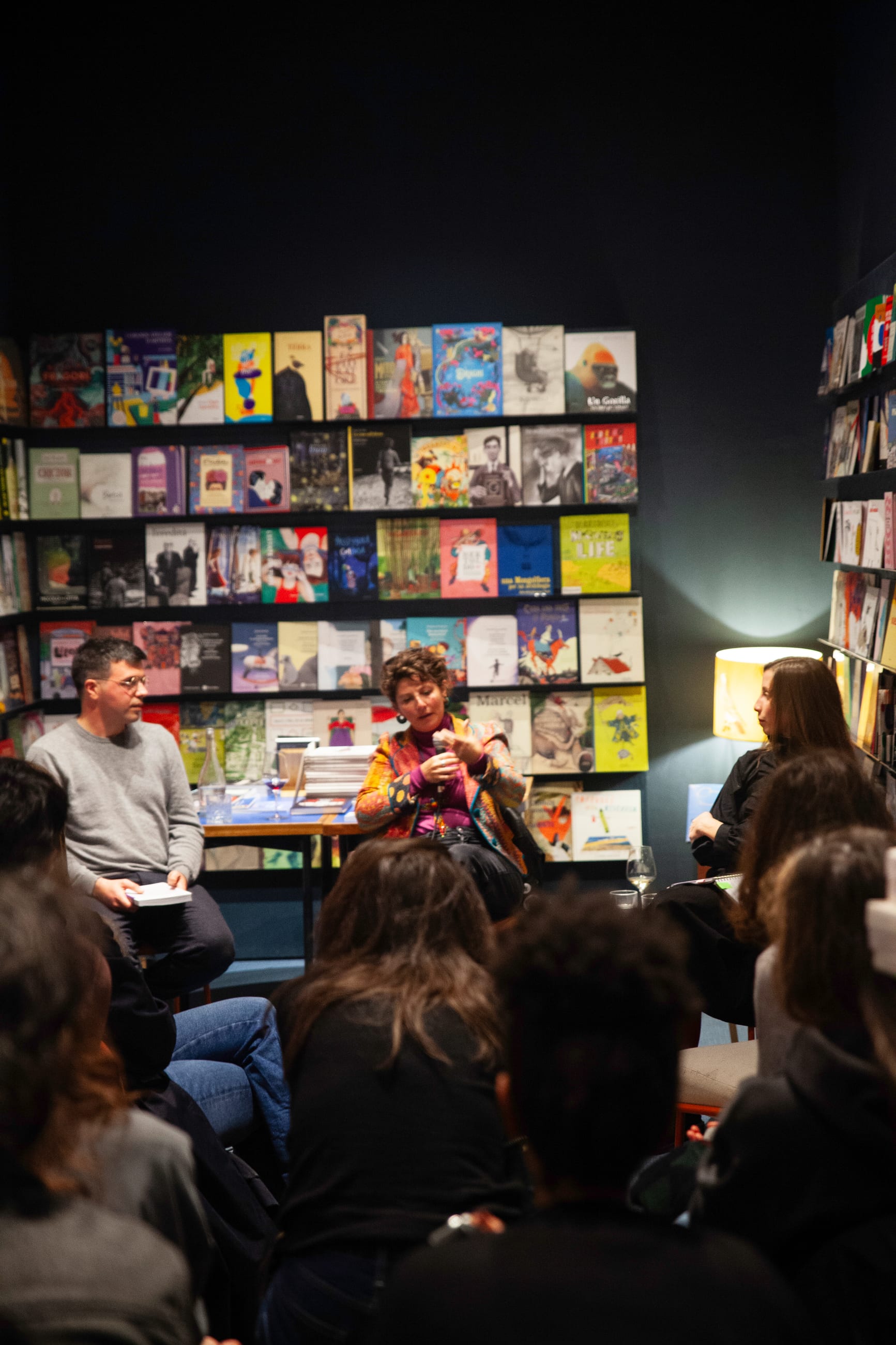 Pubblico raccolto in una libreria durante un incontro, tra scaffali di libri illuminati