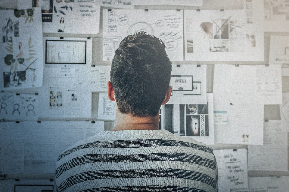 Mann von hinten blickt auf eine Wand mit zahlreichen, angehefteten Notizen und Diagrammen.