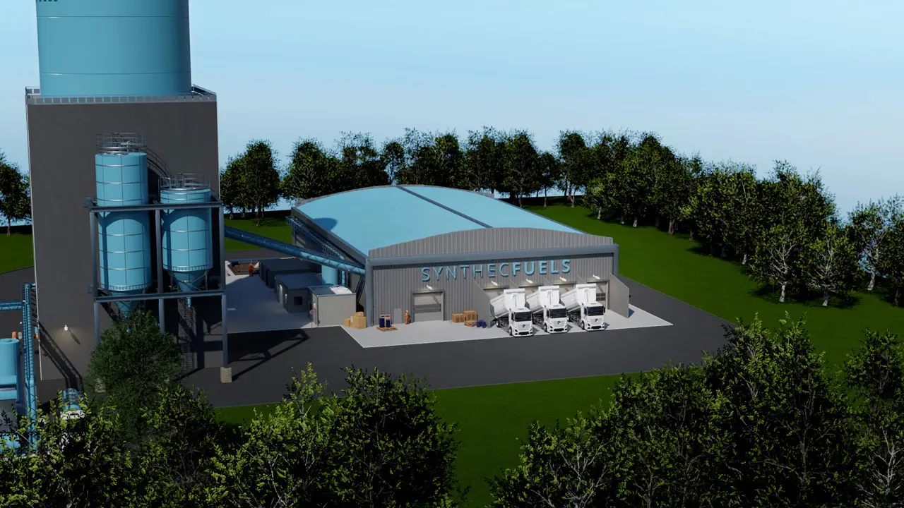 Industriegebäude mit der Aufschrift SYNTHETCFUELS, mehreren weißen LKWs und großen blauen Tanks, umgeben von Bäumen.